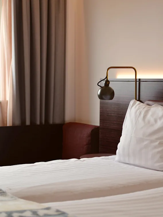 En säng med vita kuddar och en lampa på en trähuvudgavel i ett hotellrum bredvid ett fönster med gardiner.