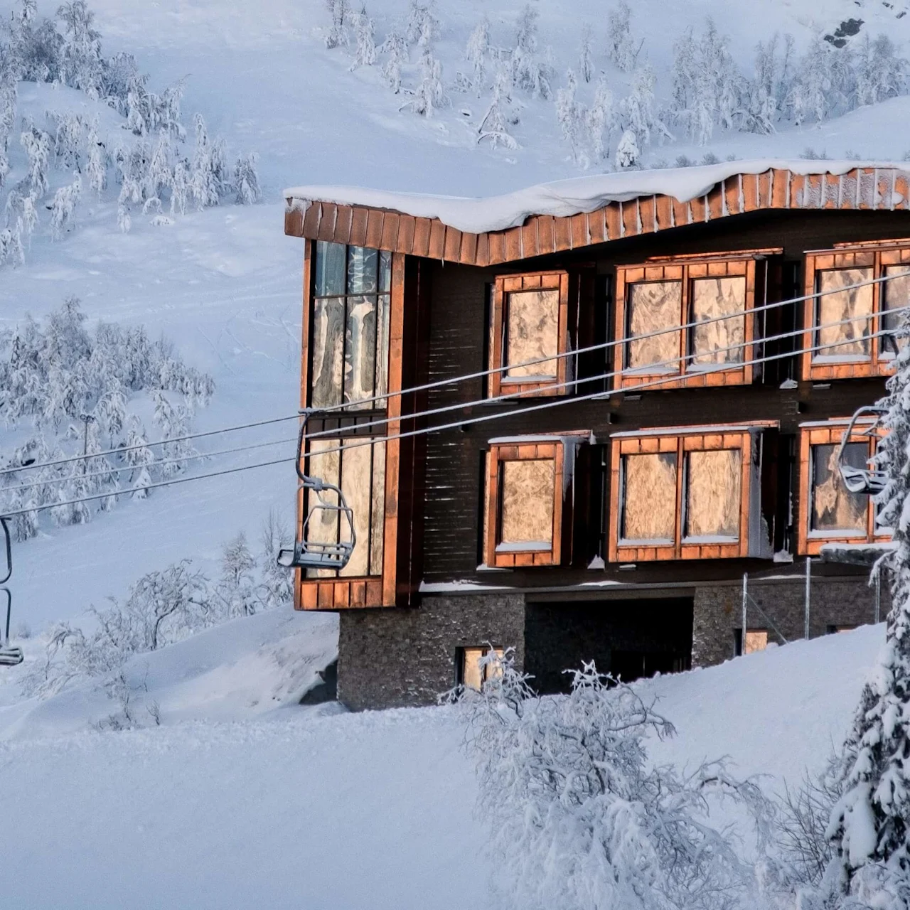 A wooden building with large windows, covered in snow, stands on a snowy mountainside. Nearby, a ski lift ascends towards snow-laden trees under a clear sky.