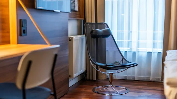 A hotel room featuring a modern chair with a mesh design and a headrest placed near a window with light curtains. Next to the chair is a wooden desk with a simple chair, and the room is fitted with wooden flooring.