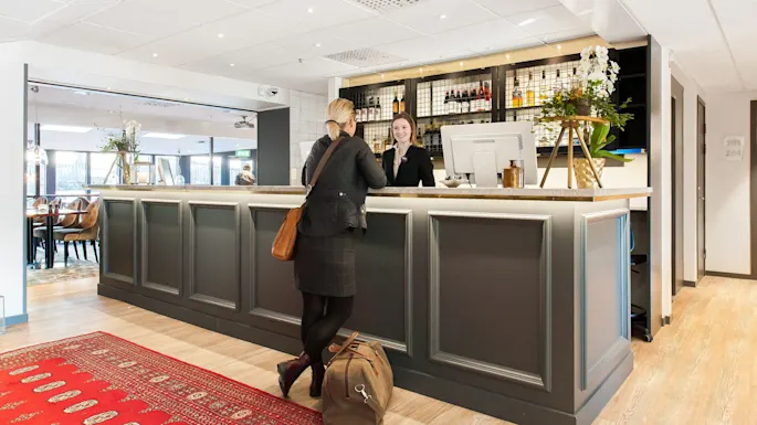 A woman checks in at a hotel reception; the smiling receptionist greets her. Warm, modern lobby with dining area in the background.