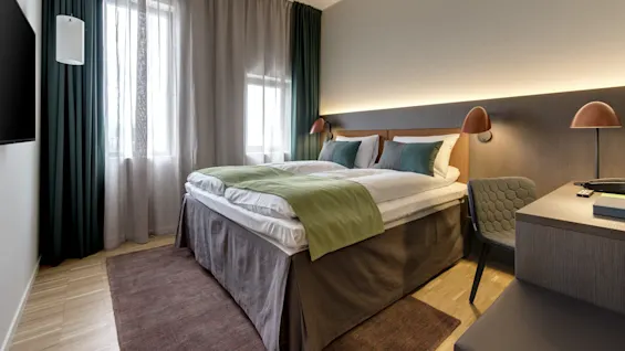 A neatly made bed with green and white linens stands in a modern hotel room. The room features wooden floors, a desk with a chair, and soft lighting.