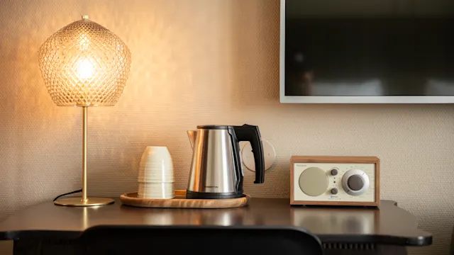 En vannkoker står på et trebord ved siden av stablede kopper og en radio. En glødende lampe lyser opp scenen ved siden av en veggmontert TV.