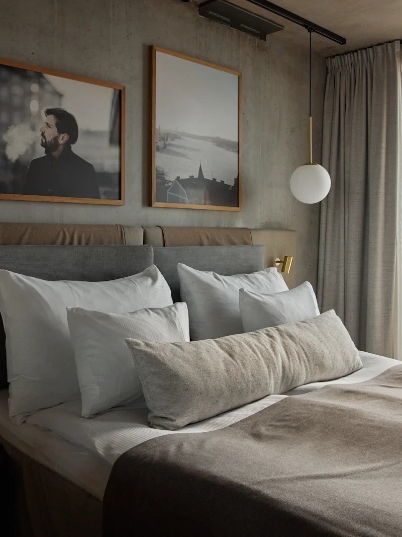 A cozy hotel room with a made bed, soft pillows, and artwork on the wall. A large window offers a city view.