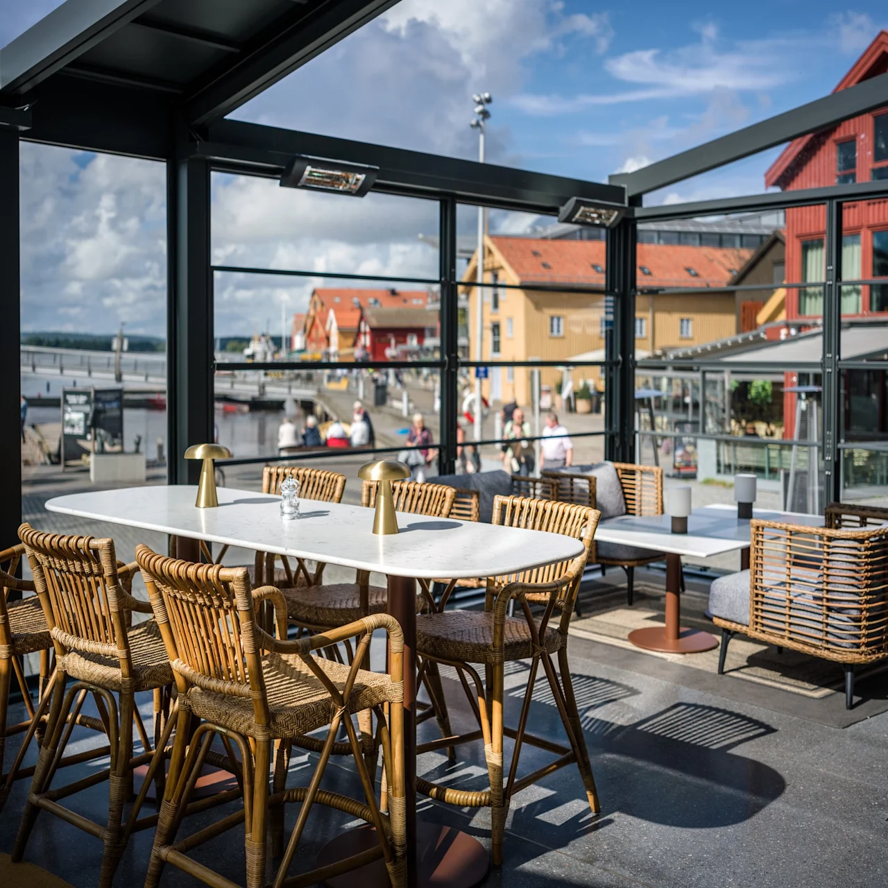 En matplats har ett vitt bord med korgstolar under ett glastak, med utsikt över en vattnkant med färgglada byggnader och fotgängare.