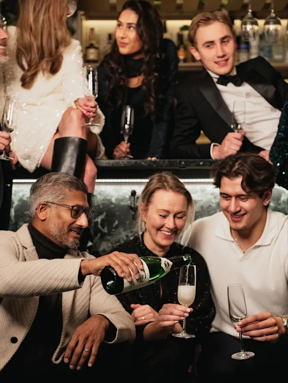 En livlig samling av vänner som njuter av champagne i en elegant miljö, klädda för fest. En man häller upp en drink med leenden omkring, fångar den glada stämningen på Clarion Hotel® Sea U.