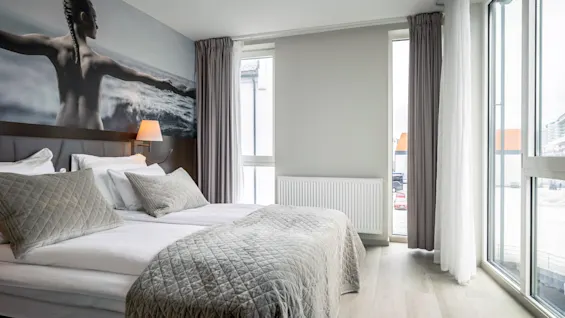En prydligt bäddad hotellsäng med grått täcke ramas in av stora fönster. En väggmålning av en person med utsträckta armar över vågor dekorerar huvudgaveln.