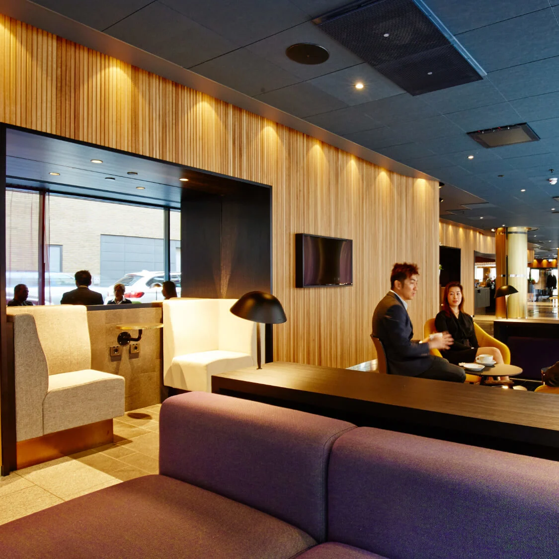 Folk sidder og snakker i en moderne lobby med trævægge og nutidige møbler. Store vinduer til højre lukker naturligt lys ind og oplyser den hyggelige atmosfære.