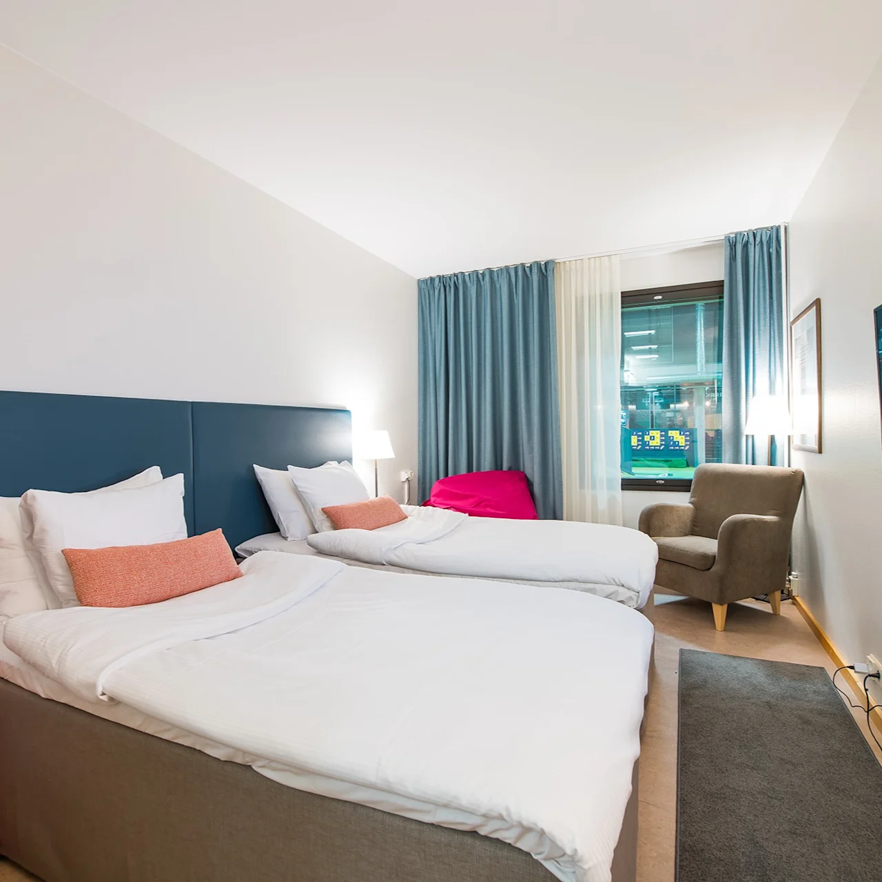 A hotel room with two neatly made beds, featuring white linens and orange cushions. A television is mounted on the wall, opposite an armchair near a window; a small red table stands beside the beds.