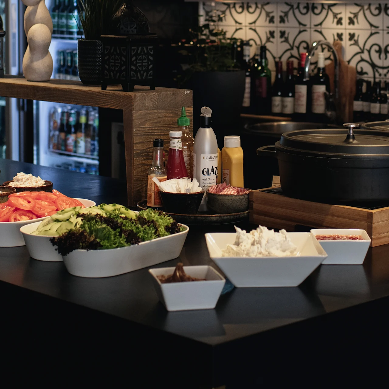 A buffet setup displays various dishes, including vegetables, rice, and sauces. Large pots rest on wooden trays. The background features patterned tiles and shelves with bottles.
