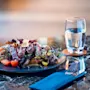 Lautasella on gourmetruokaa, jossa on viipaloitua lihaa, vihreitä ja tomaatteja marmoripöydällä vesilasillisen ja siniseen serviettiin käärityn aterimen vieressä. Sisätila.