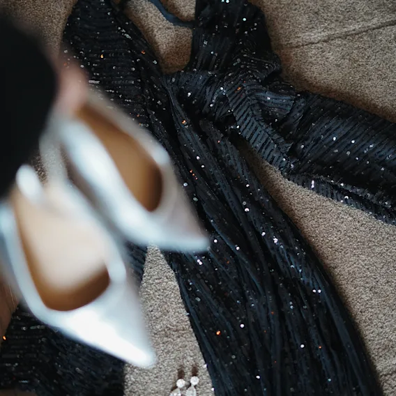 A sparkly black dress laid out on the floor with earrings, while a pair of light-colored heels is held in the foreground.