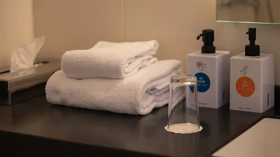 Hvite håndklær er stablet på en svart benkeplate ved siden av en eske med papirservietter, pumpeflaske merket "Go Clean Hand Soap" og en annen merket "Go Soft Body Lotion." Et glass er i nærheten.