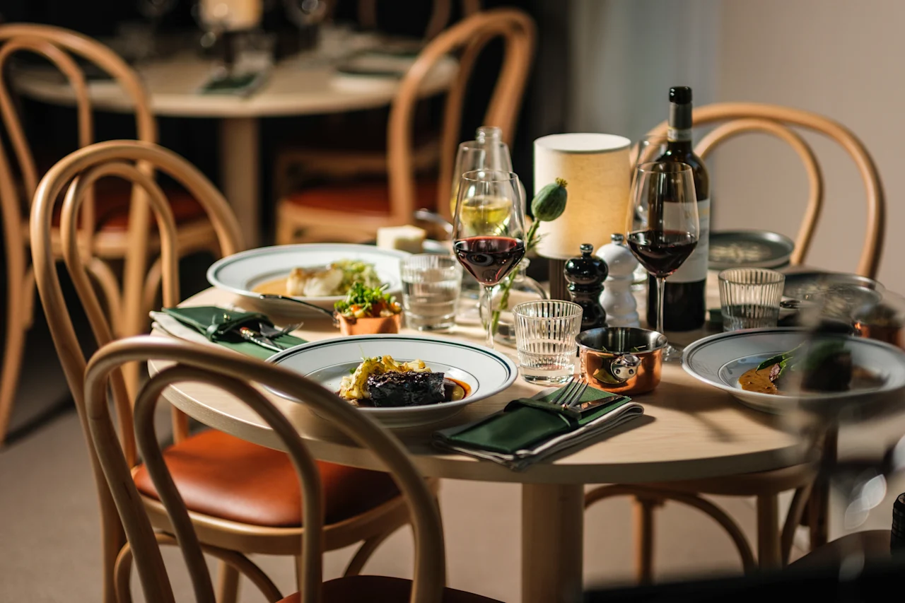 Ett runt matbord har tallrikar med gourmetmat, vinglas och en flaska, omgiven av trästolar i en varmt upplyst restaurangmiljö.