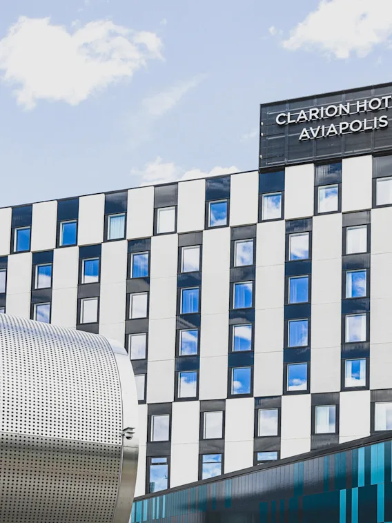 Moderni, mustavalko ruudullinen hotellirakennus seisoo sinisen taivaan alla, etualalla on sylinterimäinen metallirakenne. Kyltti lukee: "CLARION HOTEL AVIAPOLIS".