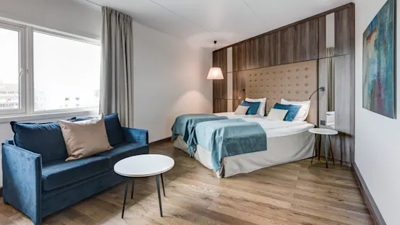 A cozy bedroom features two neatly made twin beds with blue and white bedding, a blue couch, and a small white round table. Wooden flooring and a large window provide a welcoming ambiance.