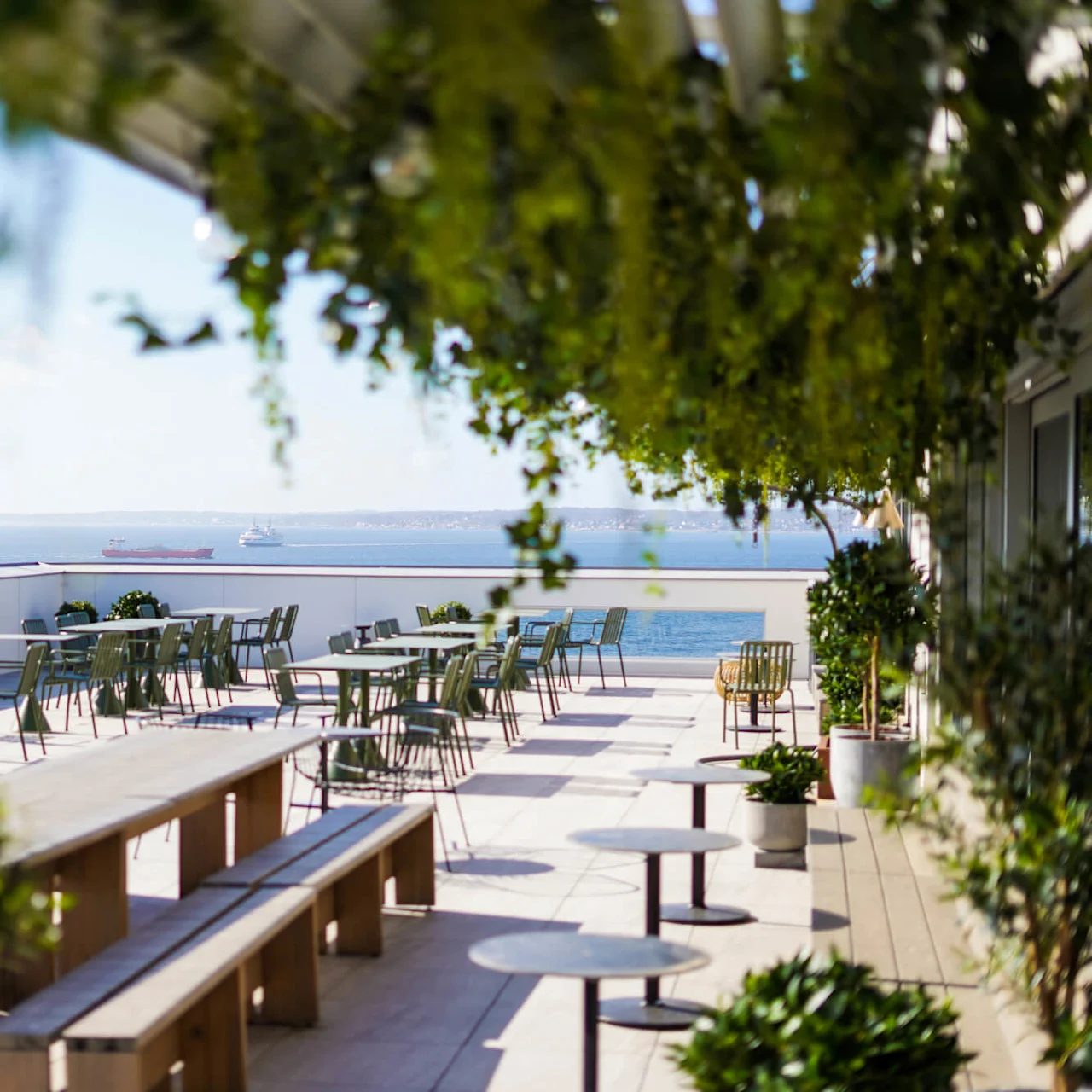 Terrasse sittegruppe med bord og stoler, omringet av løvverk, med utsikt over havet under en klarblå himmel.