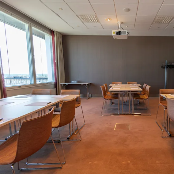 Bord och stolar är ordnade i ett konferensrum med en projektor i taket. Stora fönster släpper in naturligt ljus, och en klädhängare står i hörnet.