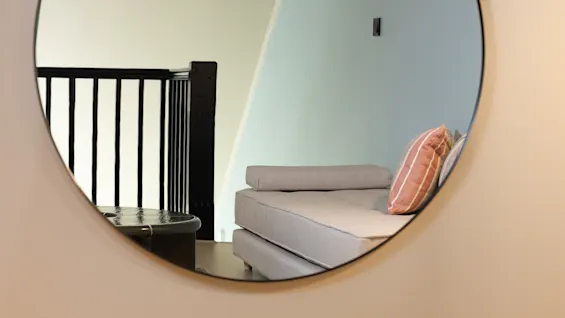 En rund spegel som reflekterar en mysig interiör med en dagbädd och en trappa, vilket skapar djup och rymd.