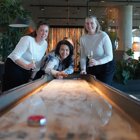 Tre personer spelar shuffleboard i en avslappnad miljö, ler och håller i drycker.