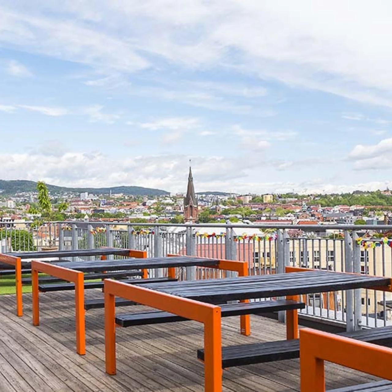 Wooden tables with orange frames stand on a rooftop terrace, overlooking a cityscape with a church spire and distant hills under a partly cloudy sky.
