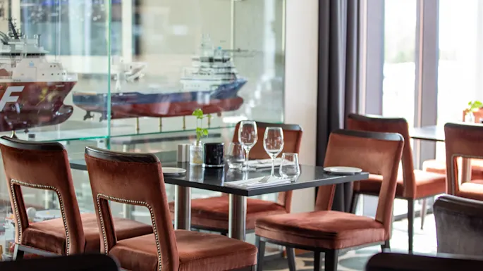 A dining table with dark surface is set with glasses, plates, and cutlery, surrounded by brown upholstered chairs. In the background, ship models are displayed in glass shelves. 