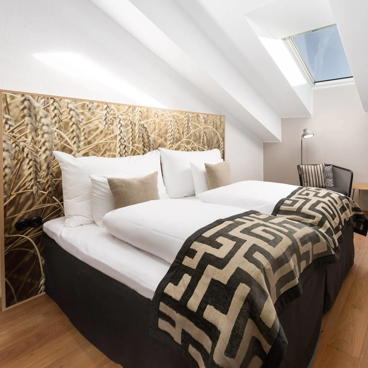 A neatly arranged attic bedroom featuring a double bed with patterned throw, a desk area with chair, under a slanted ceiling with a skylight.
