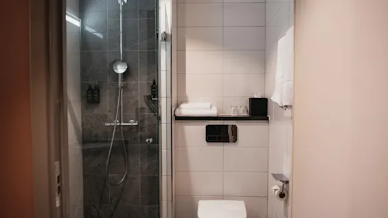 Et rent og moderne hotellbad med en romslig dusj med mørke fliser og et toalettområde med friske håndklær.
