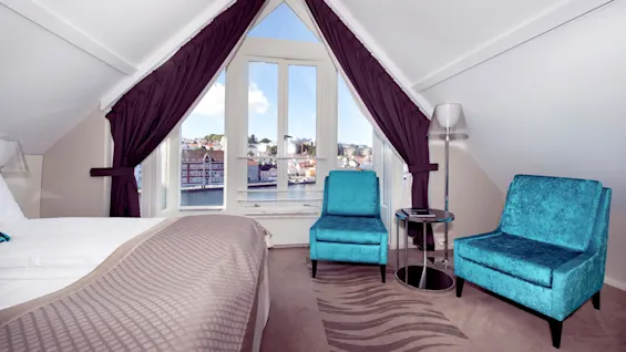 A well-lit hotel room with a bed and two turquoise chairs, large windows overlooking a waterway and European-style buildings.