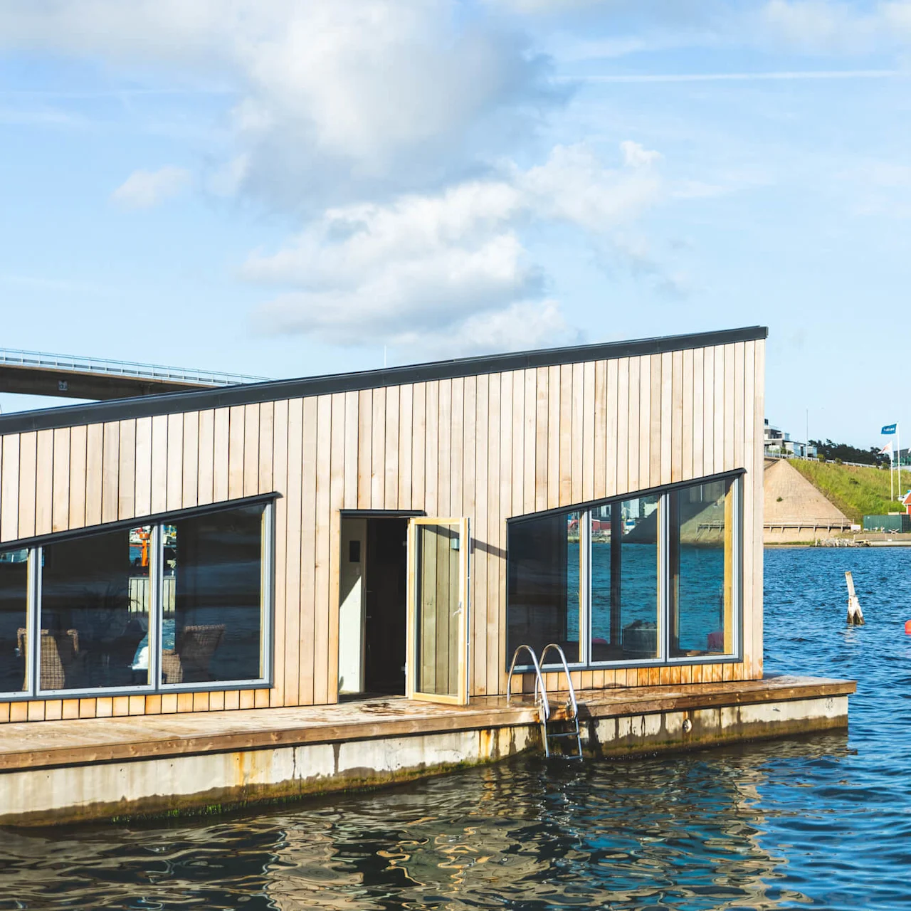 A wooden houseboat rests on calm water, with windows reflecting the blue sky, surrounded by a coastal town with red cabins, flags, and modern buildings.