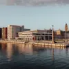 Moderne bygningskompleks ved en havnepromenade med urban arkitektur, herunder en karakteristisk murstenstårnstruktur, under en klar himmel i dagslys.