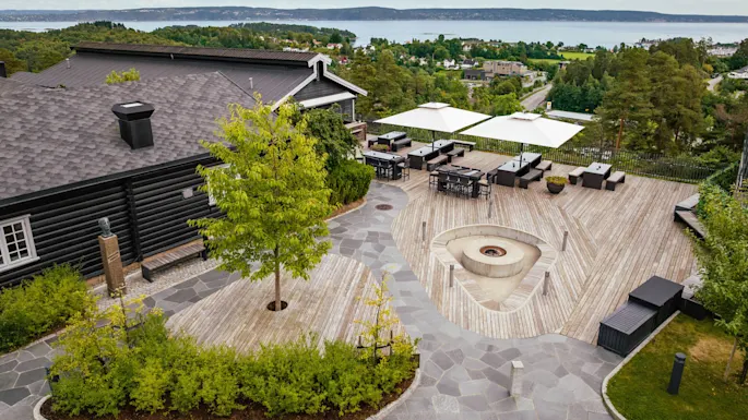 A spacious wooden deck features modern outdoor furniture and a central fire pit. Surrounded by lush greenery, it overlooks a scenic landscape with distant water and rolling hills.