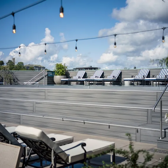 Solstoler med stripete puter er arrangert på en takterrasse, omgitt av potteplanter. Lysslynger henger over, med utsikt over bygninger og en skyet himmel i bakgrunnen.