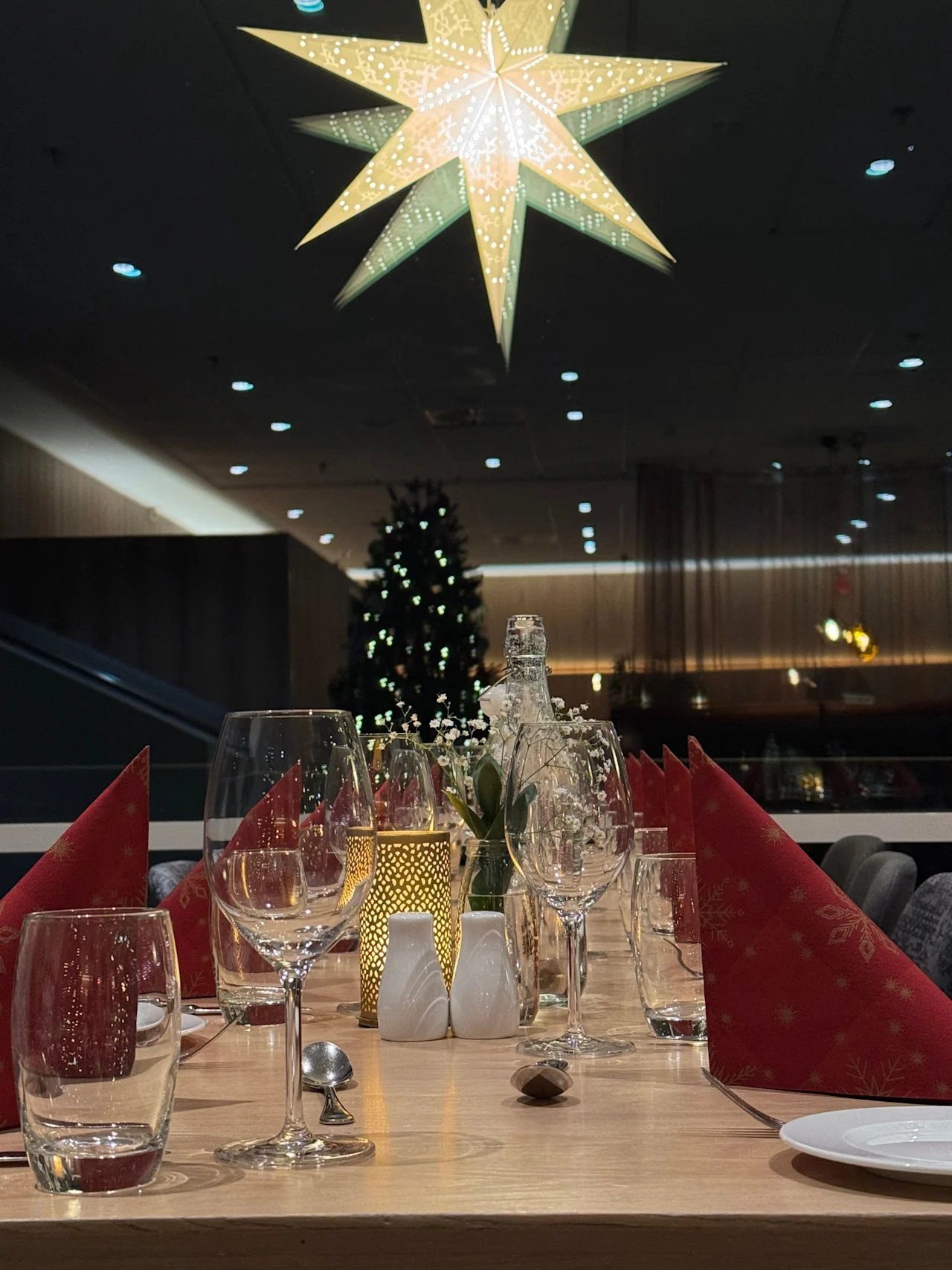 Et bord er elegant dekket med vinglass, røde servietter og en flaske. Over henger en lysende stjerne. I bakgrunnen er et juletre synlig i et svakt opplyst rom.