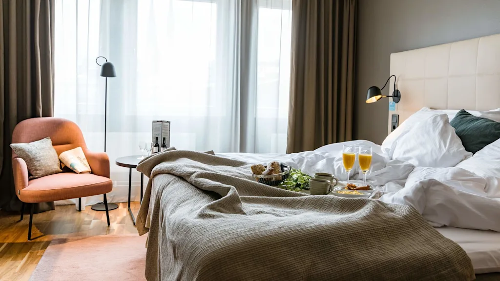 Seng med frokostbrett, inkludert croissanter og appelsinjuice, i et godt opplyst, koselig hotellrom med beige sengegavl, brune gardiner og en oransje lenestol ved vinduet.