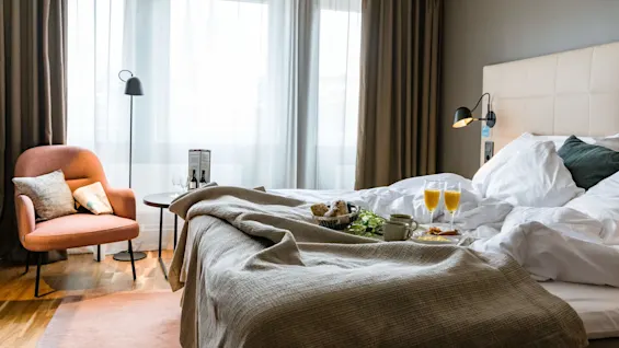 Bed with breakfast tray, containing croissants and orange juice, in a well-lit, cozy hotel room with a beige headboard, brown curtains, and an orange armchair by the window.