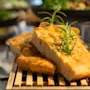 Skiver af brød toppet med rosmarin hviler på en træbakke, med en sløret baggrund af forskellige retter på en buffet.