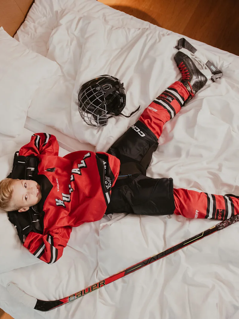 En ung hockeyspiller i rødt og svart utstyr ligger spredt ut på en seng, med hjelm ved siden av seg, hockeykølle på gulvet og hvitt sengetøy i et rom med treverk.