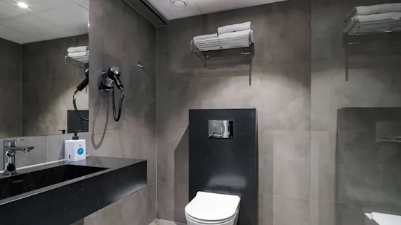 Badeværelse med en moderne vask og vandhane, som spejles i et spejl, en vægmonteret hårtørrer, toilet med metaltryk, og hylde med foldede håndklæder. Grå flisebelagte vægge skaber en smart stemning.