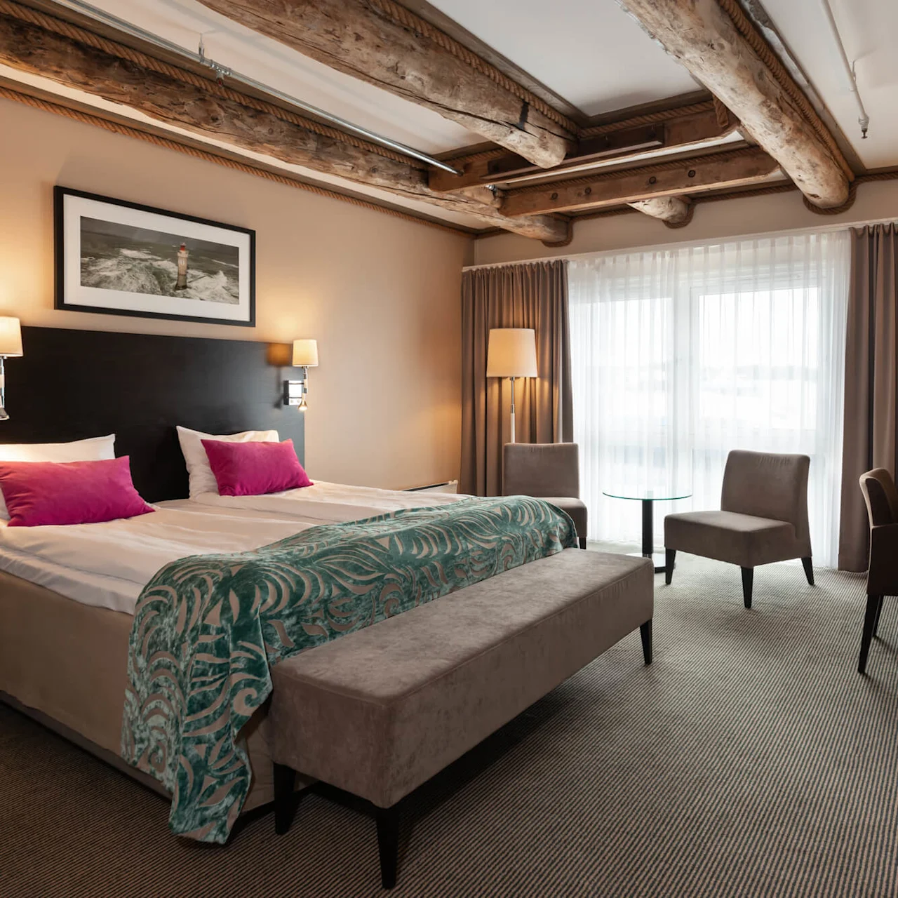 A neatly arranged hotel room features a large bed with pink pillows, a wooden desk, two chairs, and a patterned teal blanket, situated in a modern setting with wooden beam details.
