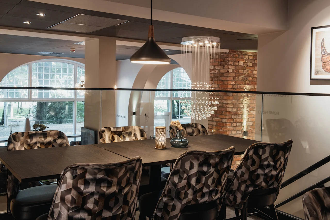 Et stilfuldt spiseområde med et langt mørkt bord, mønstrede stole og store buede vinduer. Synlig tekst: HOME HOTEL.