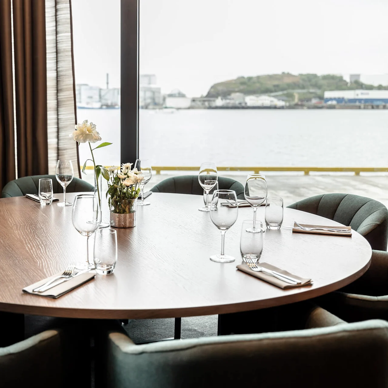 Round table set with wine glasses and napkins, surrounded by upholstered chairs, in a dining room with large windows overlooking a waterfront and industrial buildings in the distance.
