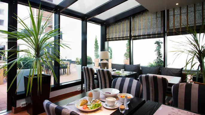 Frokostmat ordnet på et bord med stripete stoler, inne i et lyst rom med glassvegger og potteplanter, med utsikt over en uteplass med flere sitteplasser.