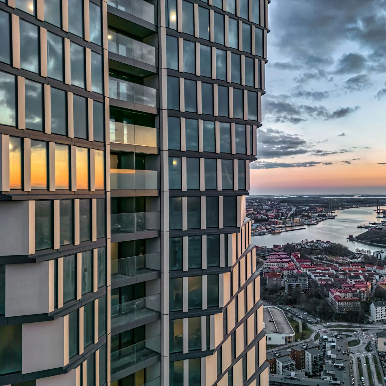 En höghusbyggnad med reflekterande fönster står vid en flod i solnedgången, med utsikt över en stadsmiljö med rödklädda tak och avlägsna industribyggnader under en delvis molnig himmel.