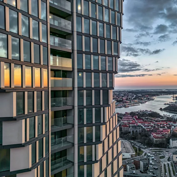 En höghusbyggnad med reflekterande fönster står vid en flod i solnedgången, med utsikt över en stadsmiljö med rödklädda tak och avlägsna industribyggnader under en delvis molnig himmel.
