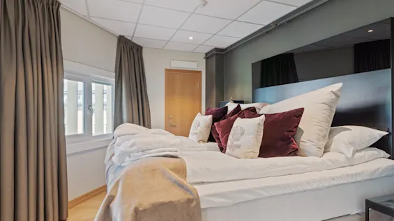 A bed with white linens and burgundy pillows is unmade in a modern bedroom. Thick curtains adorn the windows, and a wooden door is closed, enhancing the cozy atmosphere.