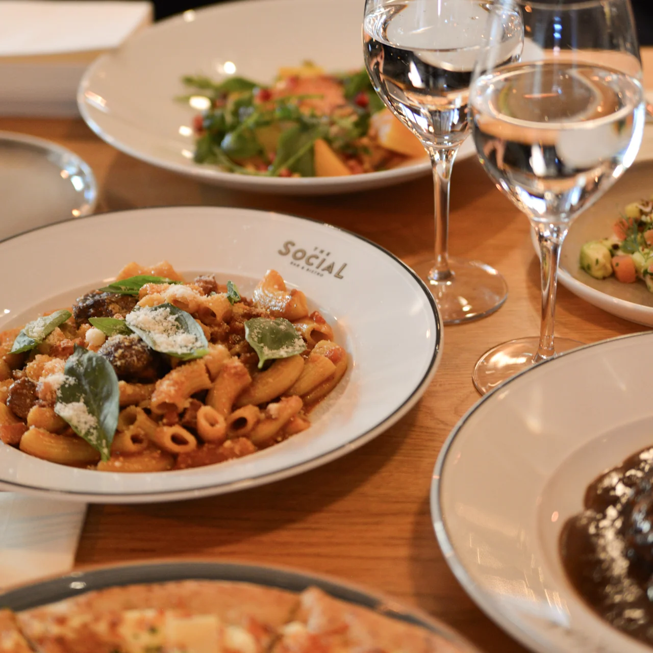 En pastaret pyntet med urter ligger på en tallerken mærket "SOCIAL". Omgivende retter og to vinglas antyder en restaurantindstilling med forskellige retter.