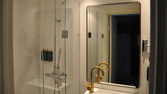A sleek bathroom features a glass shower enclosure with chrome fixtures and mounted black toiletries. A gold faucet is reflected in a large square mirror under recessed lighting.