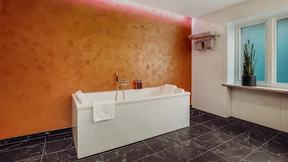 A modern bathtub against a textured orange wall, with ambient lighting and a potted plant near the window.