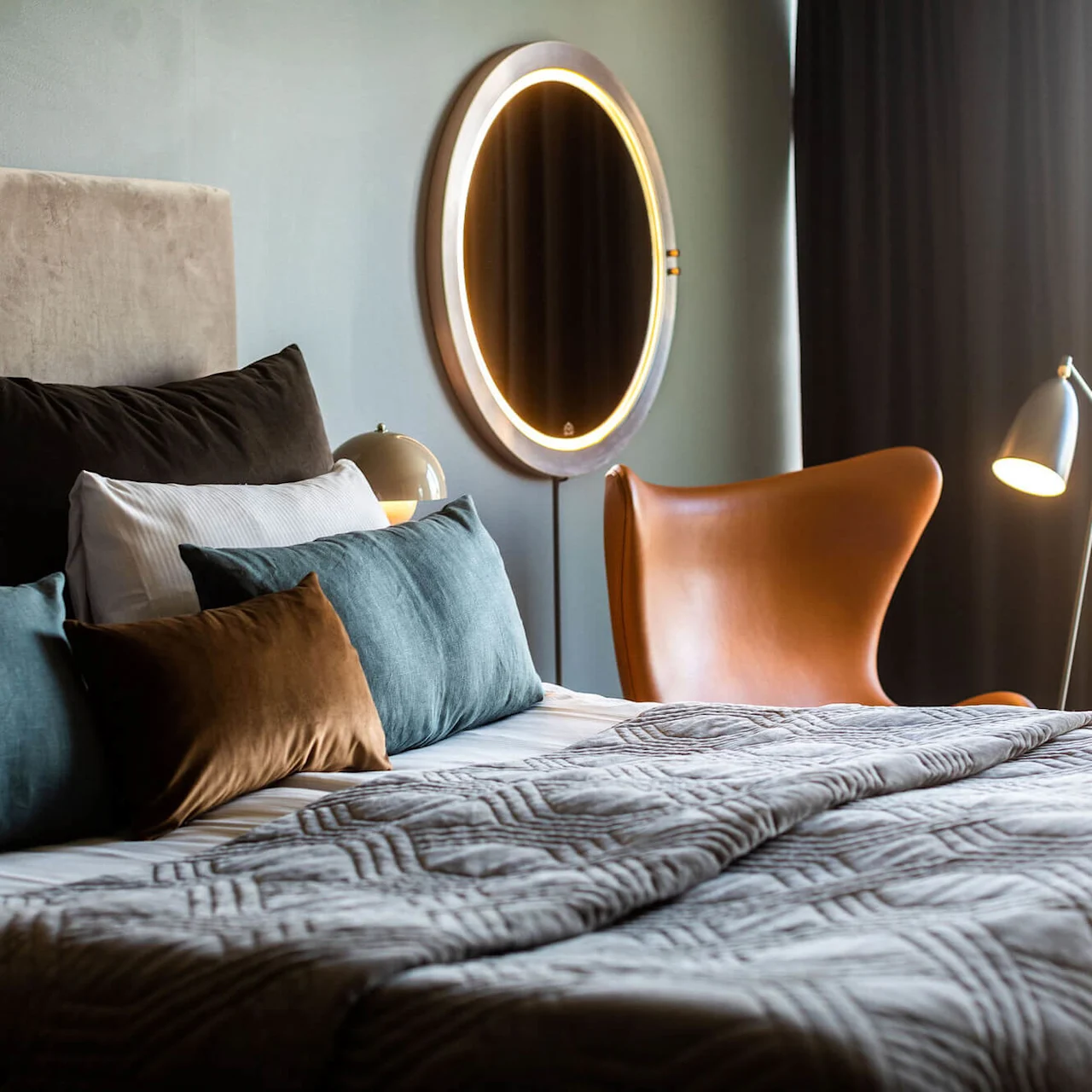 Säng prydd med flerfärgade kuddar, täckt av ett täcke, placerad nära en brun stol under en spegel med lampor och bredvid en lampa; i ett mjukt belyst, modernt rum.