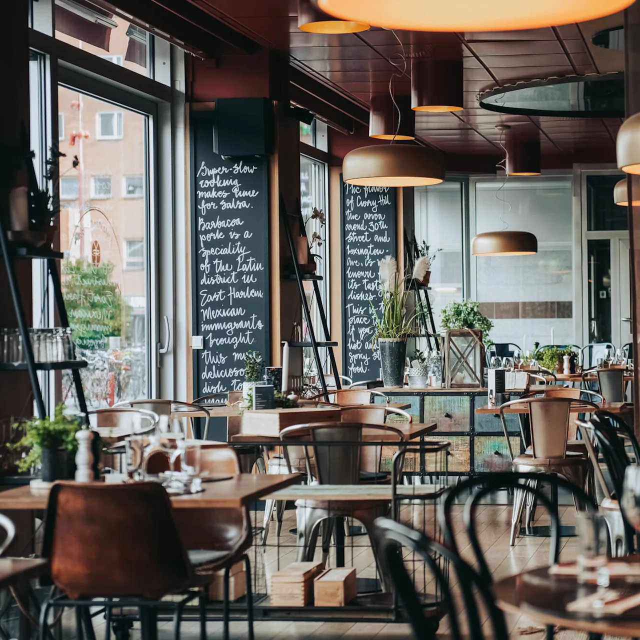 Elegant restaurant with wooden tables set for dining, surrounded by large windows and blackboards displaying handwritten text. Soft lighting fixtures hang from the ceiling, creating a cozy ambiance. 